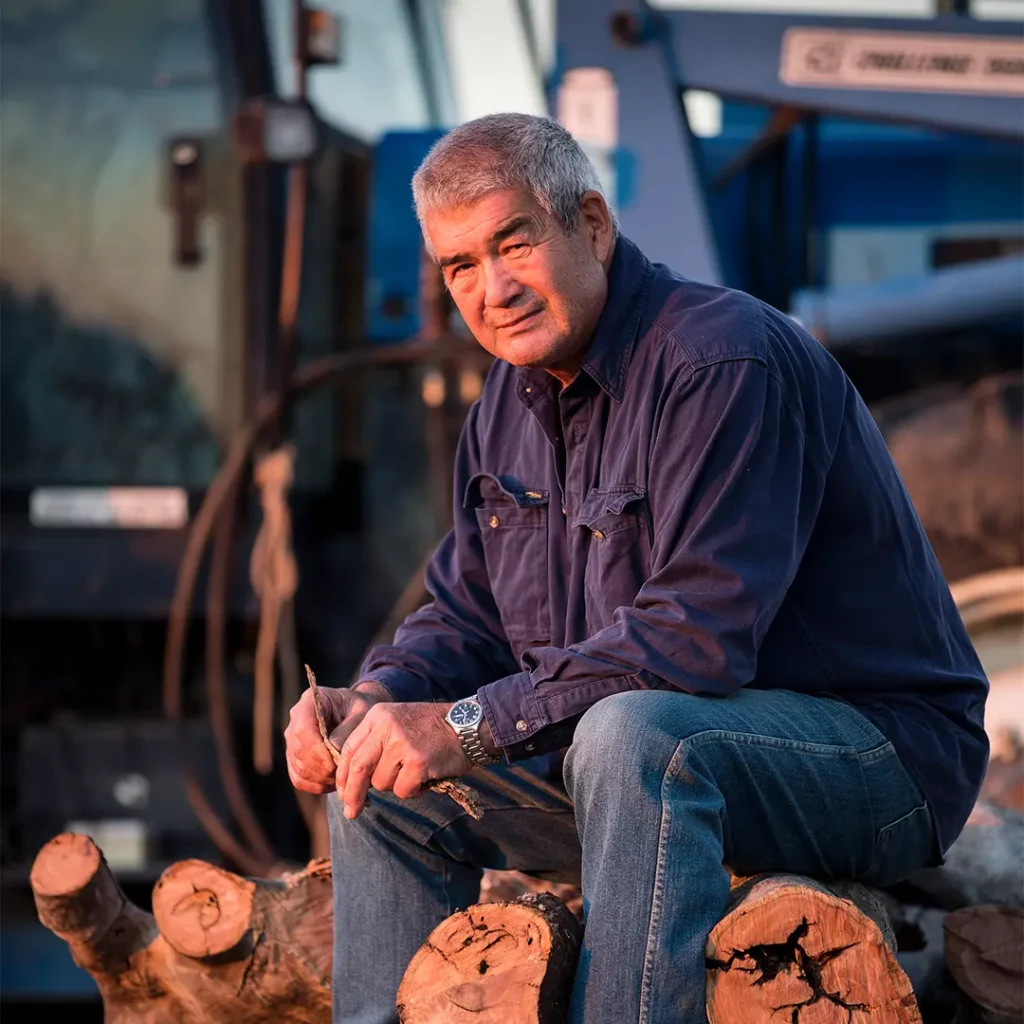 A wheat/sheep farmer John Harper sitting on a cut tree log in the early morning sun, his machinery behind him.